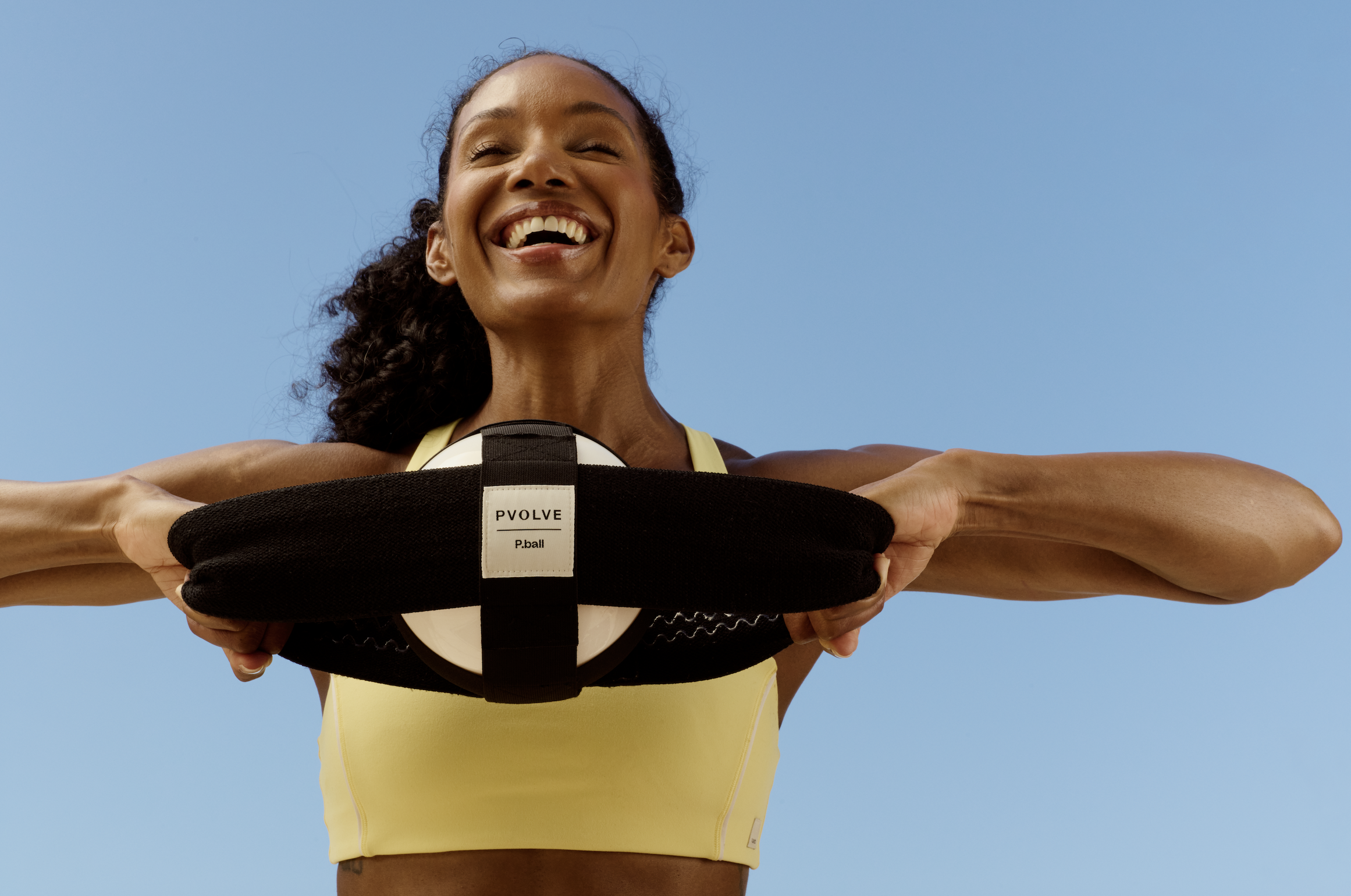 A smiling woman in a yellow sports top holds a Pvolve P.ball exercise tool against a blue sky.