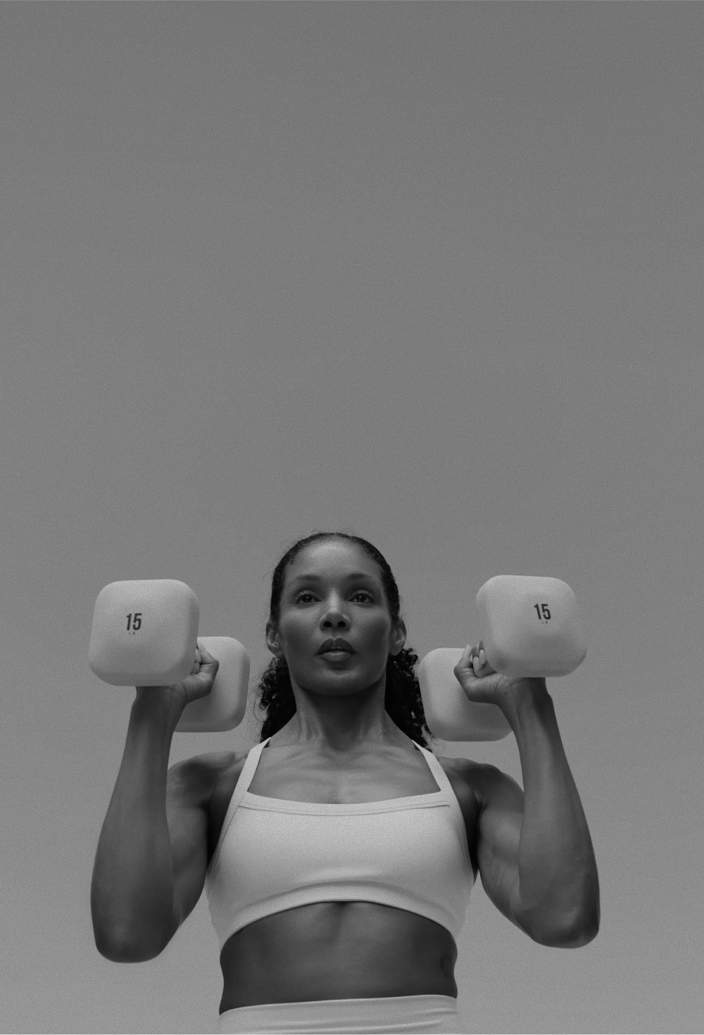 A low-angle, black and white photo of a woman lifting two 15-pound dumbbells.