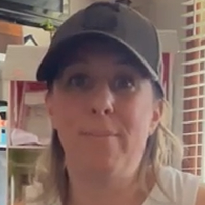 A close-up of a woman with blonde hair wearing a dark baseball cap and a white shirt.