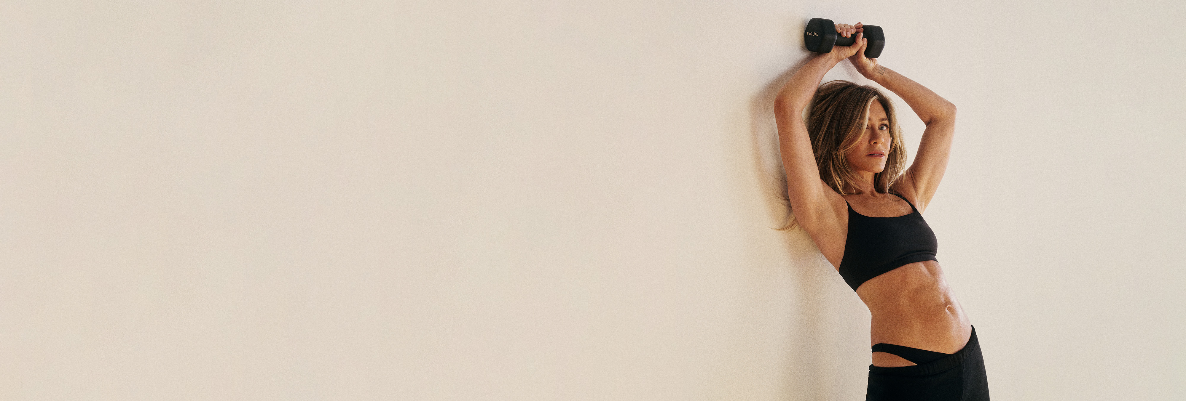 A woman in black workout clothes holds a dumbbell above her head, leaning against a plain wall.