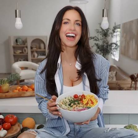 A smiling woman with dark hair sits on a counter holding a large bowl of salad.