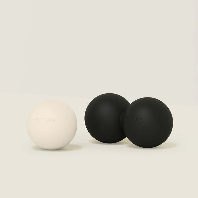 A single off-white Pvolve massage ball sits next to a black double massage ball against a cream background.