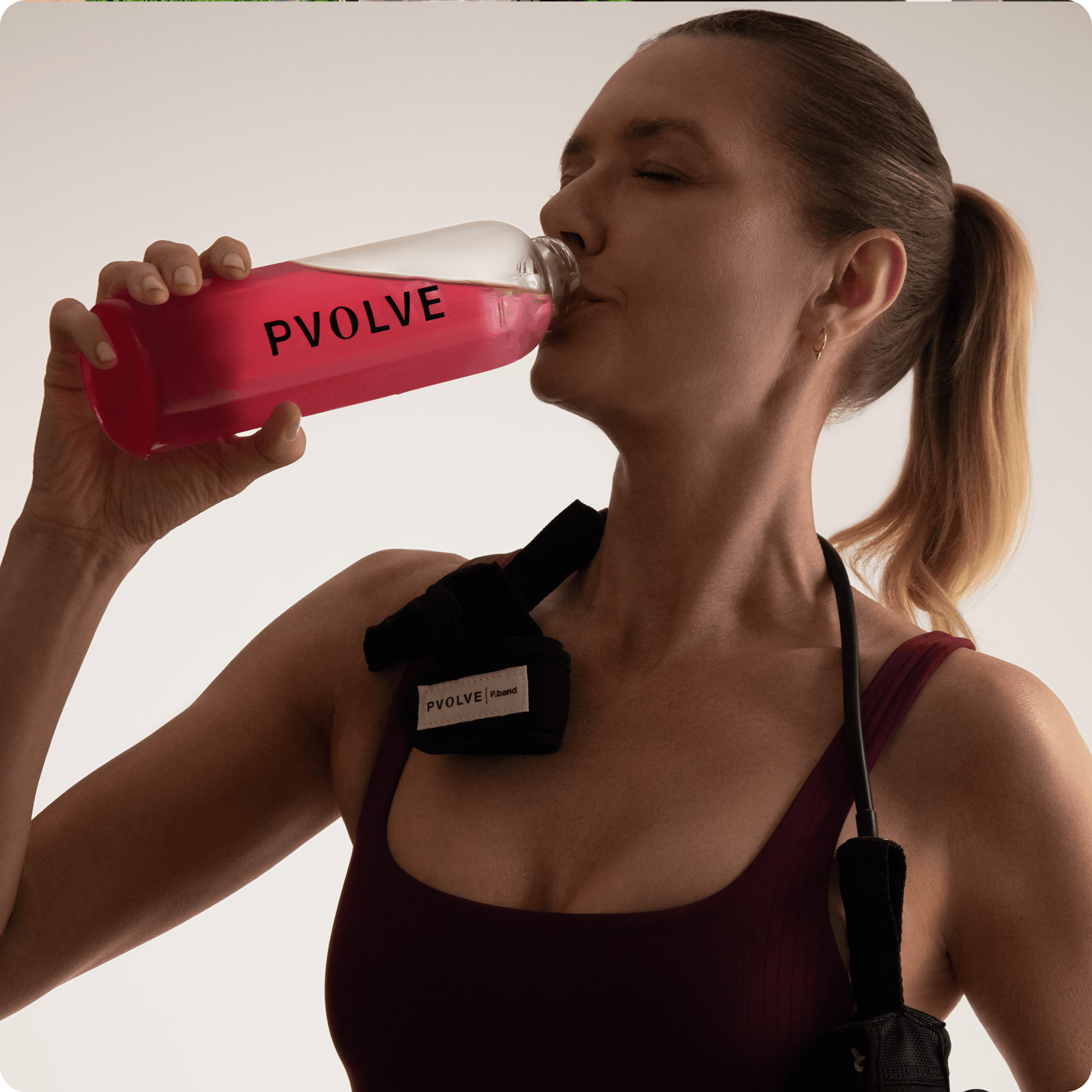 A woman in workout gear with a P.band over her shoulder drinks from a Pvolve branded water bottle.
