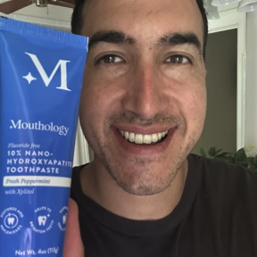 Person smiling and holding a blue toothpaste tube labeled 'Mouthology.'