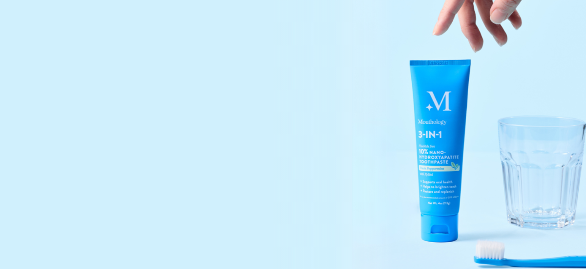 A hand reaching for a tube of toothpaste with a toothbrush and glass on a light blue background.