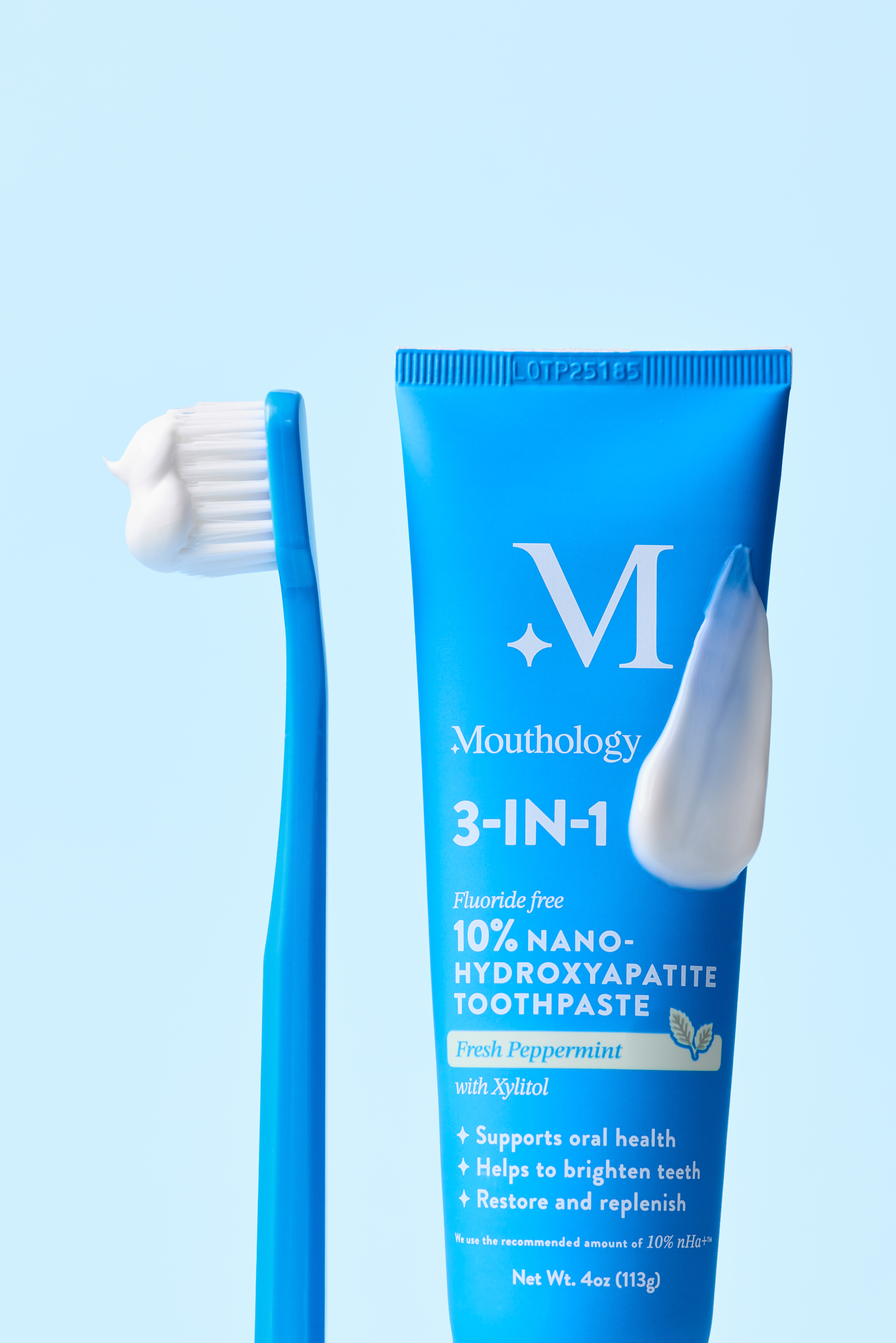 A blue tube of Mouthology toothpaste stands next to a blue toothbrush with white toothpaste on it.