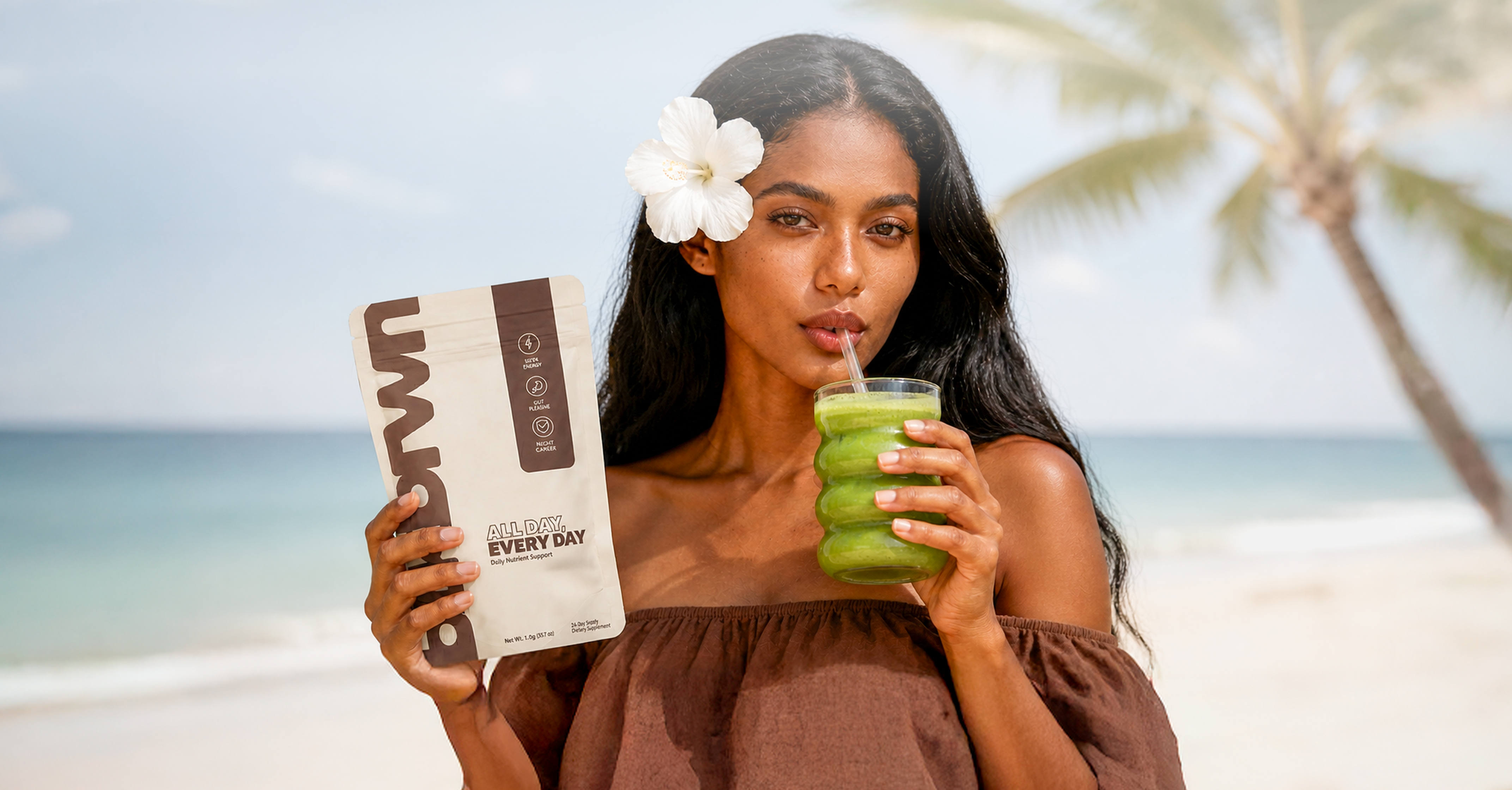 A woman on a beach holds a product pouch while drinking a green smoothie.