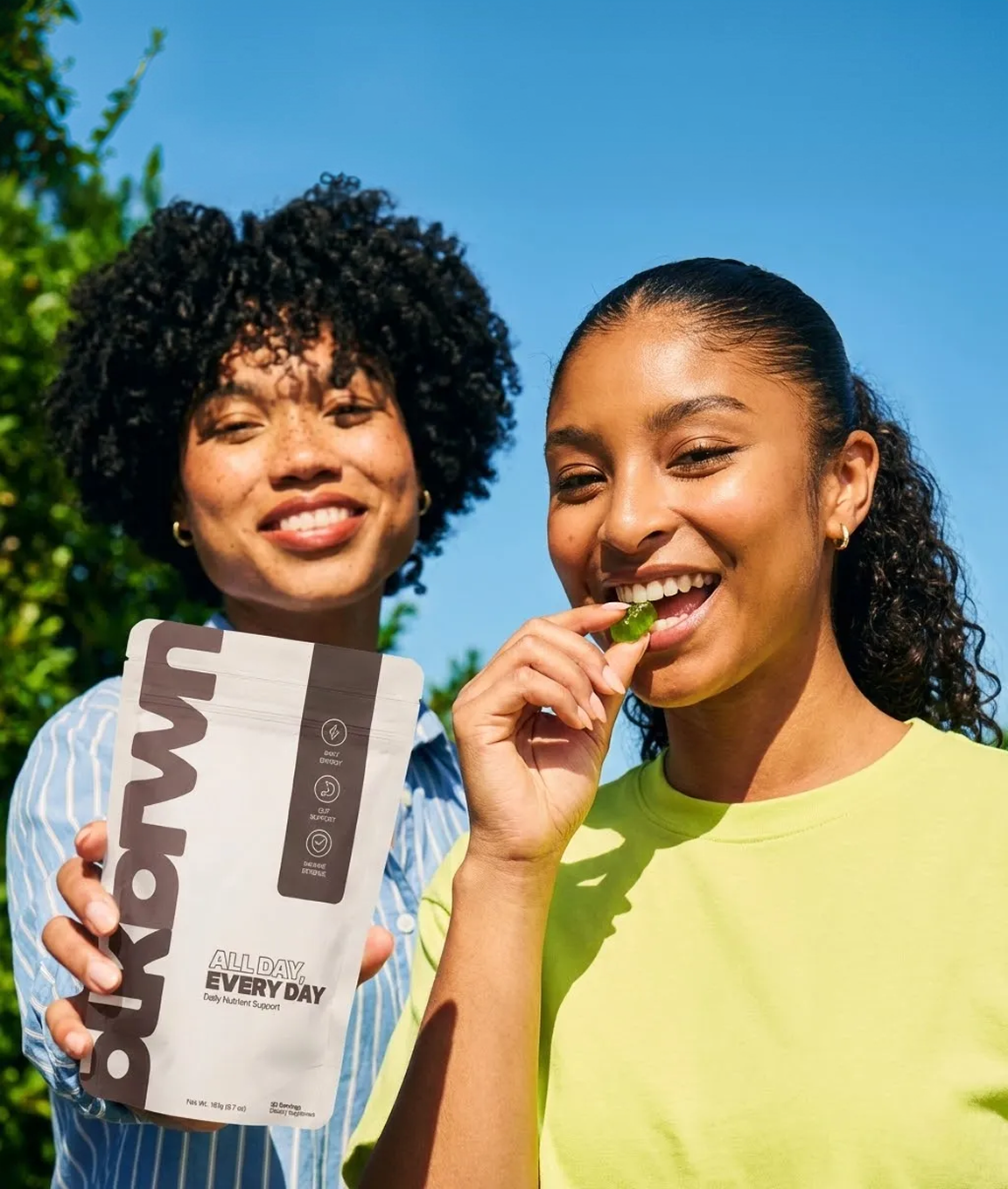 Two women smiling outdoors, one holding a bag of supplements while the other eats a green gummy.
