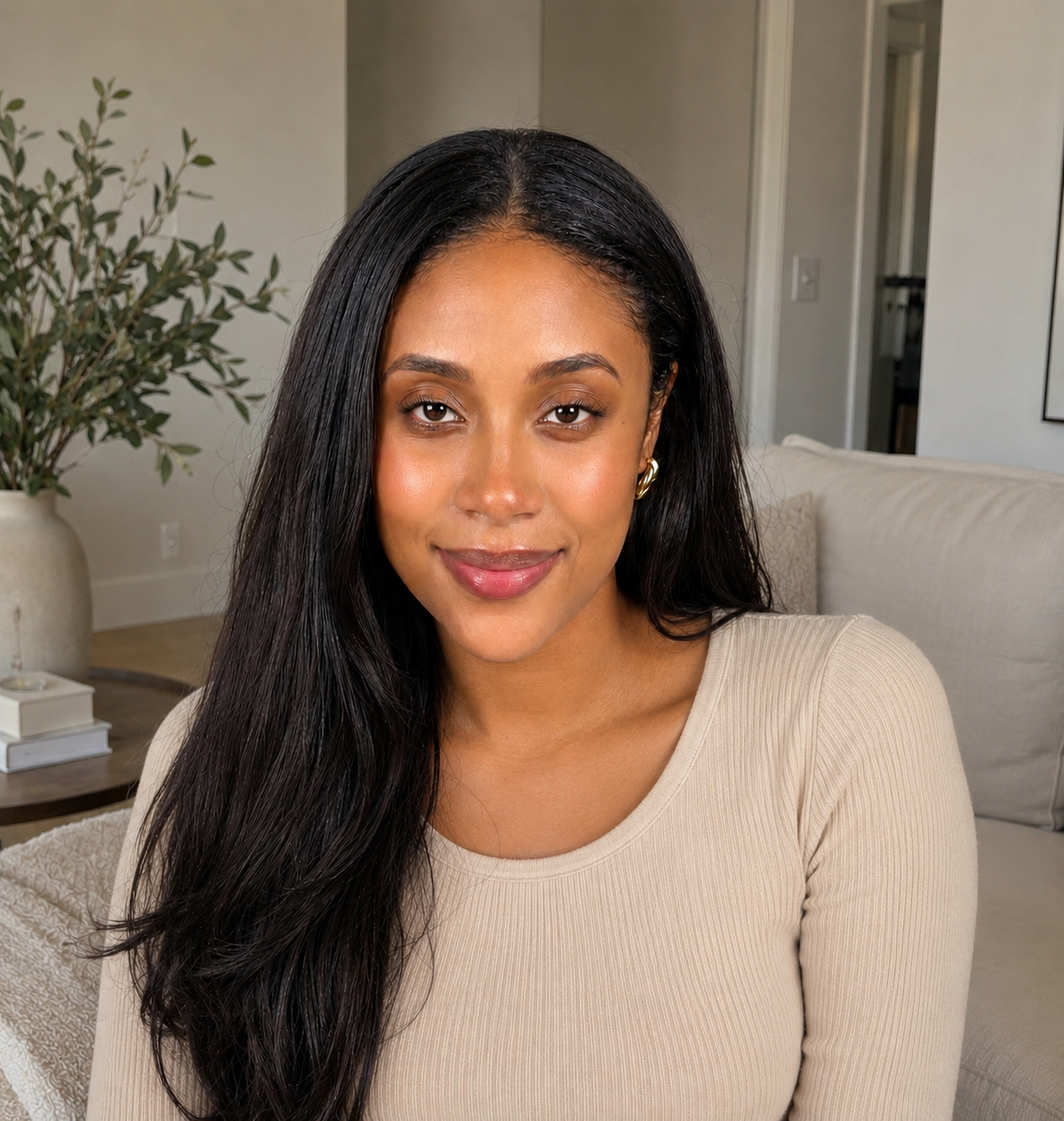 A woman with long, dark hair and a beige ribbed top smiles at the camera in a living room.