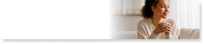 A woman sits on a couch, smiling and holding a mug while looking out a window.
