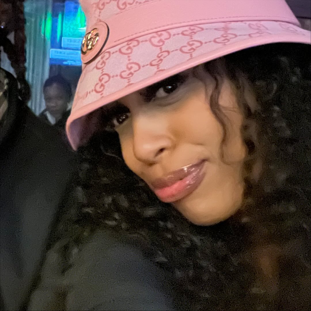A close-up of a person with long, dark curly hair wearing a pink bucket hat and smiling.