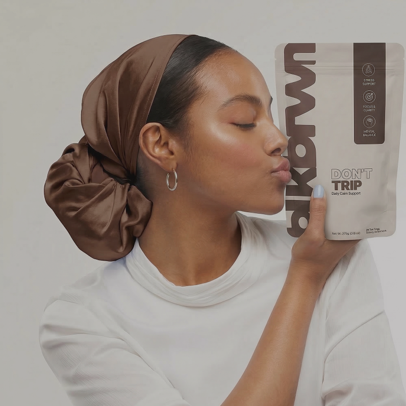 A woman with a brown silk head wrap holds a white pouch to her lips with her eyes closed.