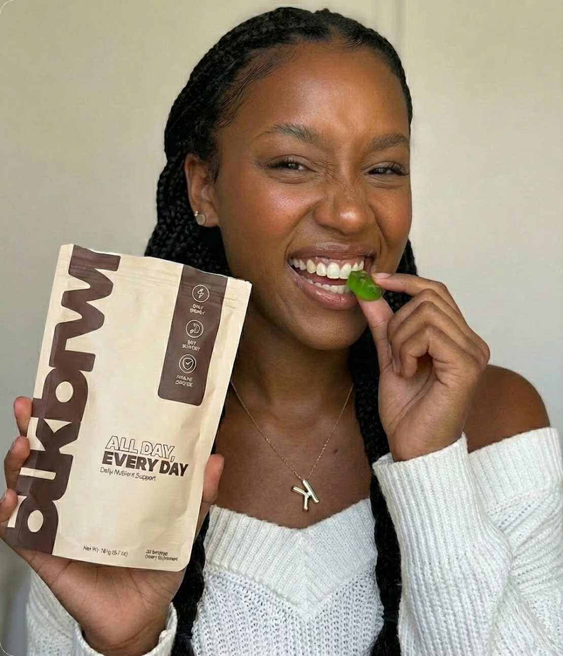 A smiling woman with braids holds a pouch of 'All Day, Every Day' and eats a green gummy.