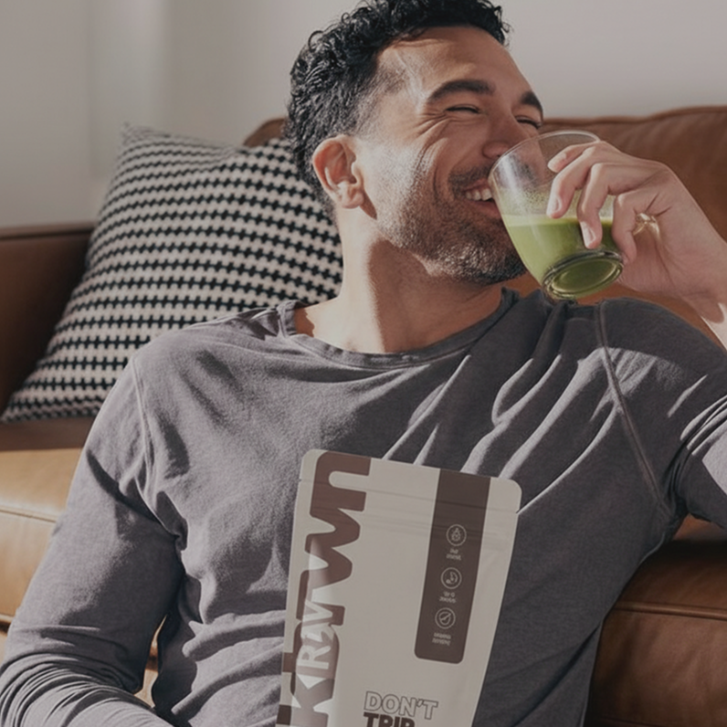 A man smiles while sitting on a couch, drinking a green beverage and holding a pouch.