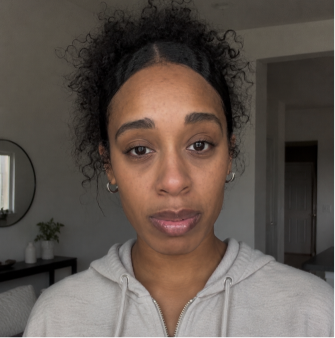 A woman with dark, curly hair pulled back in a light-colored hoodie looks directly at the camera.