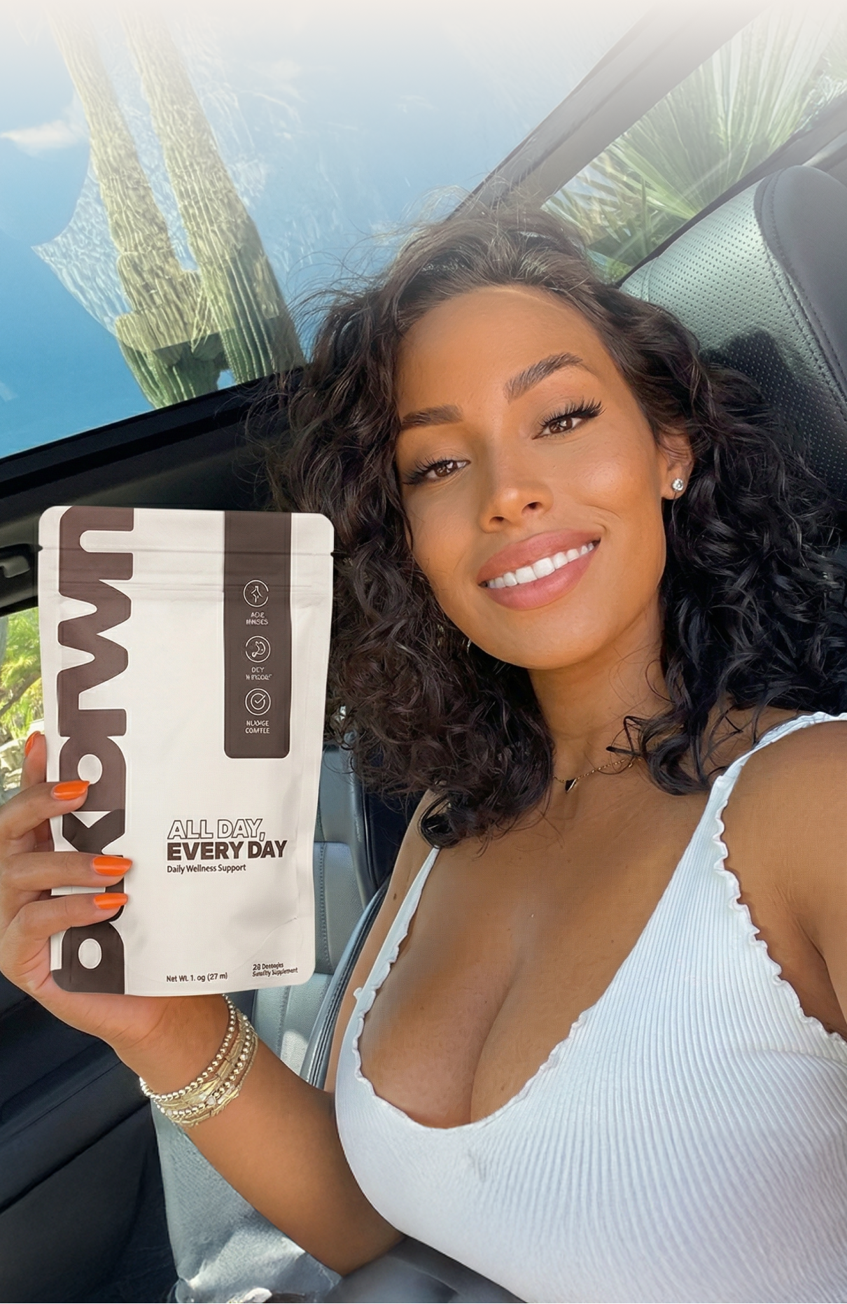 A woman with curly hair smiles in a car, holding a white pouch of wellness supplements.