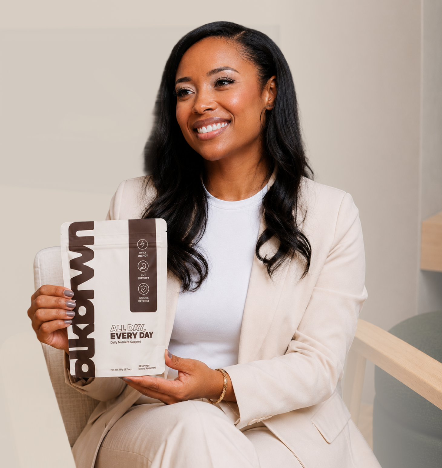 A smiling woman in a beige suit sits in a chair, holding a pouch of daily nutrient support.