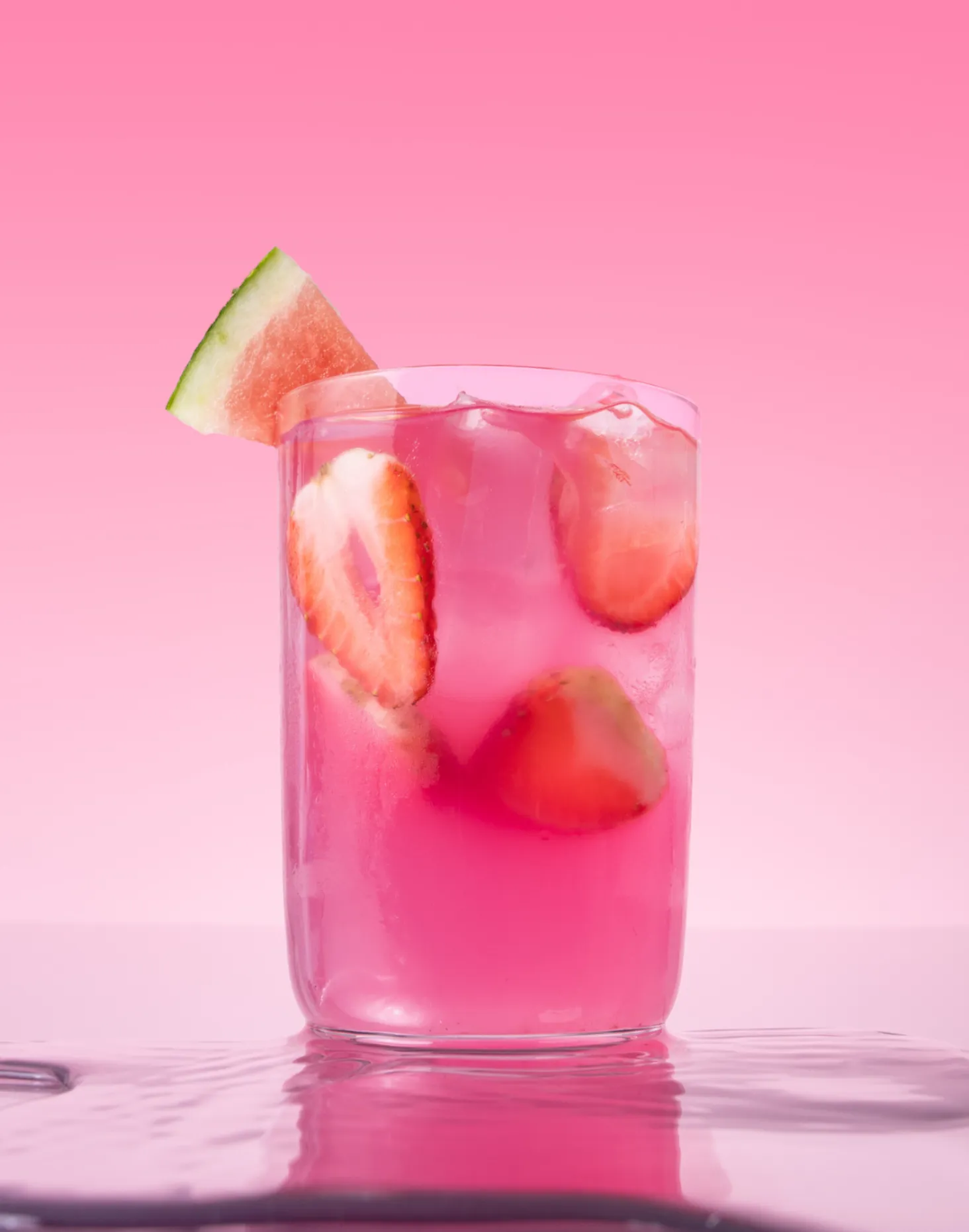 A tall glass filled with pink juice, ice, and sliced strawberries is garnished with a watermelon wedge. The background is a smooth gradient of light pink.