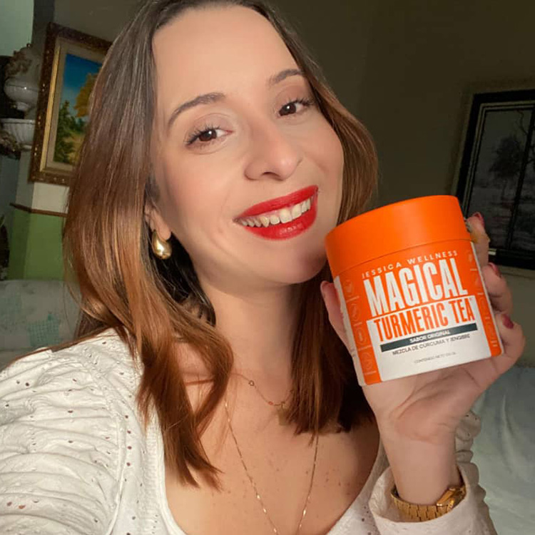 A smiling woman with red lipstick holds up a container of Jessica Wellness Magical Turmeric Tea.
