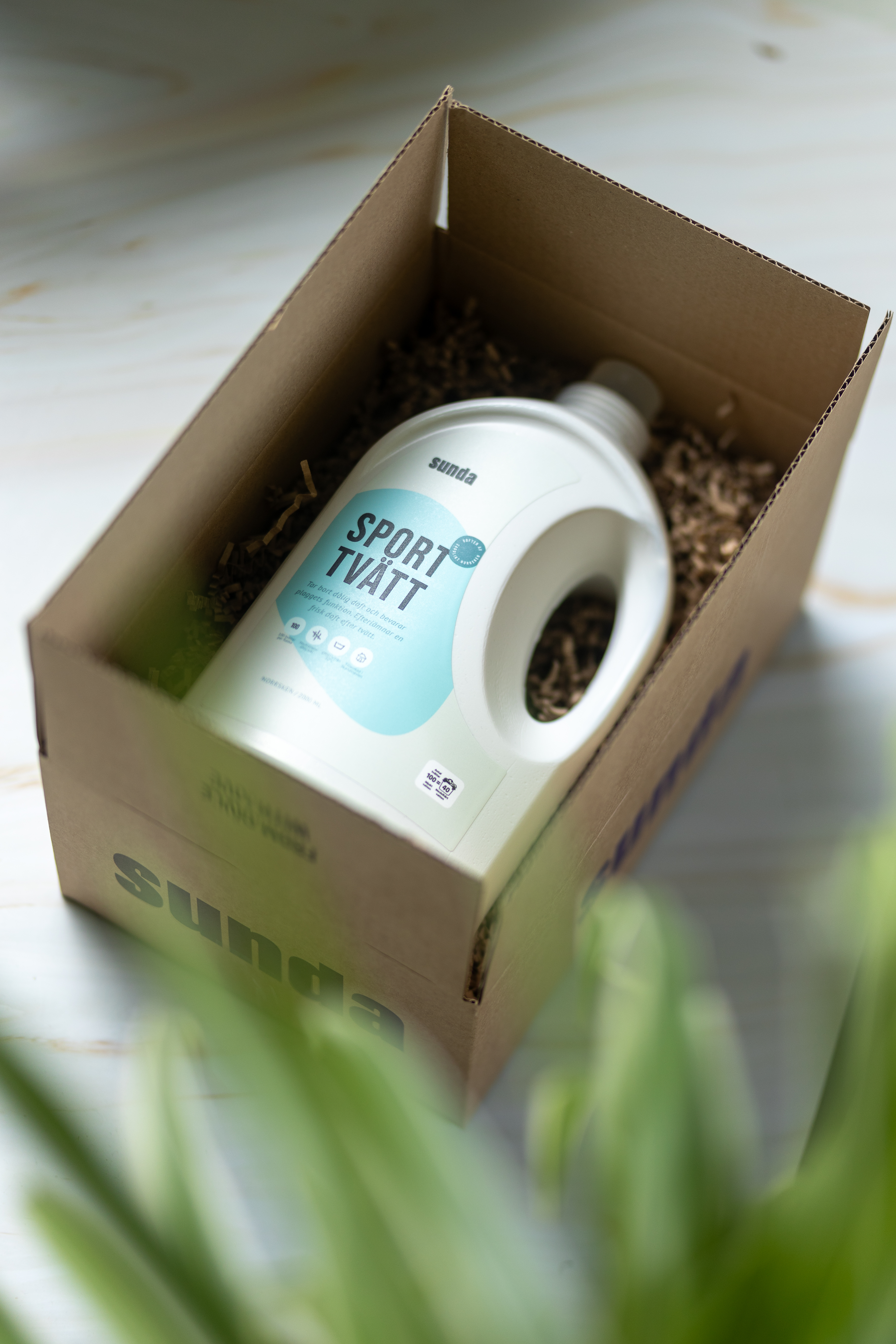 A bottle of detergent labeled 'Sport Tvätt' in a cardboard box with shredded paper.