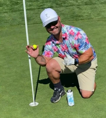 Man in floral shirt and cap kneeling on golf course with ball near hole.