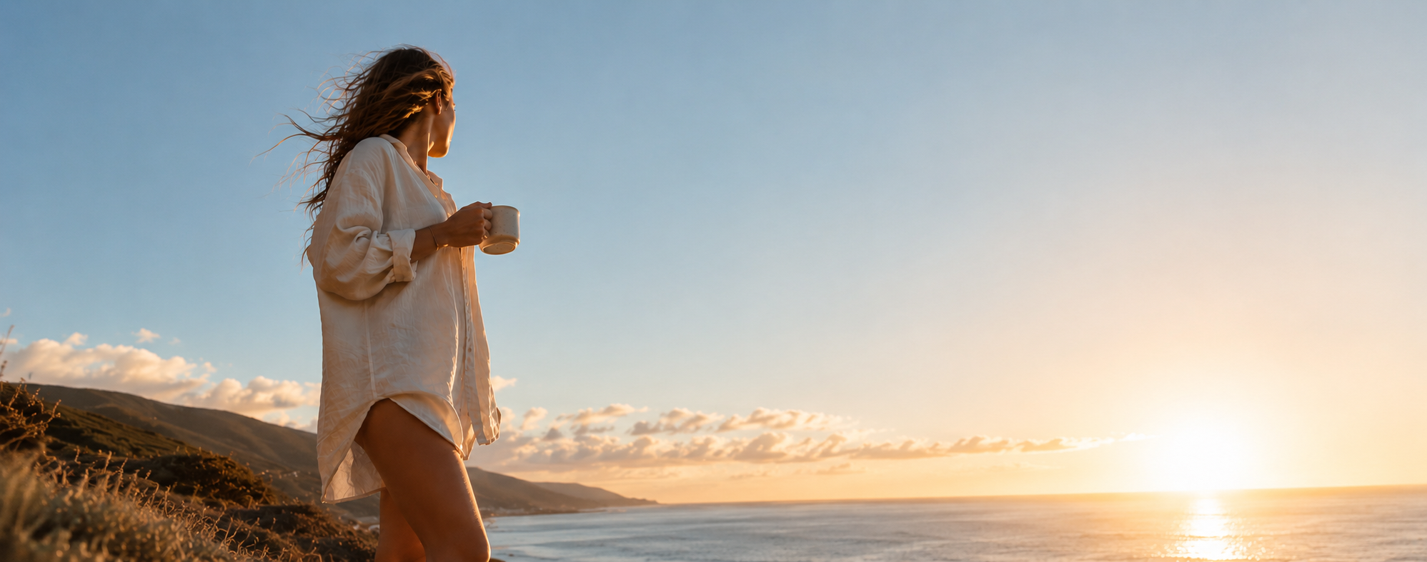 A woman holds a mug on a hillside, looking out at the ocean during sunset.