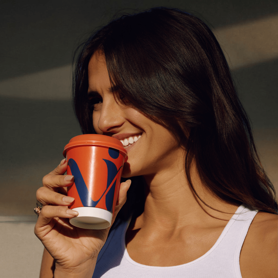 A smiling woman with long dark hair holds an orange and blue coffee cup to her lips.