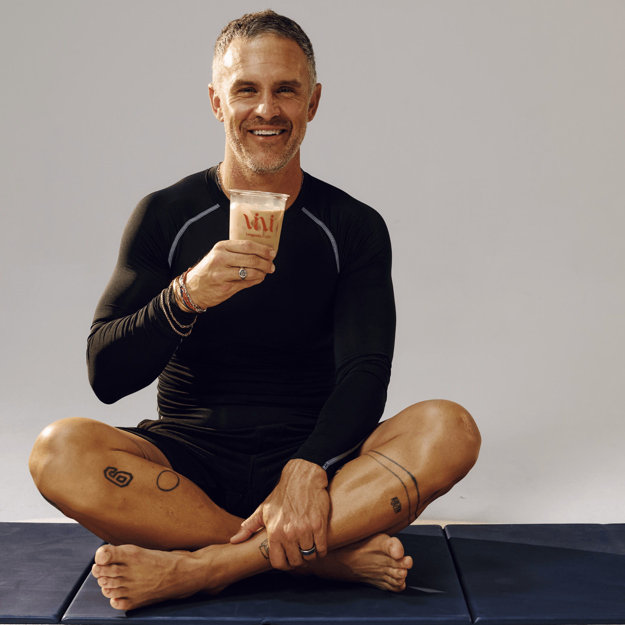 A smiling man in black athletic wear sits cross-legged on a mat, holding a drink.