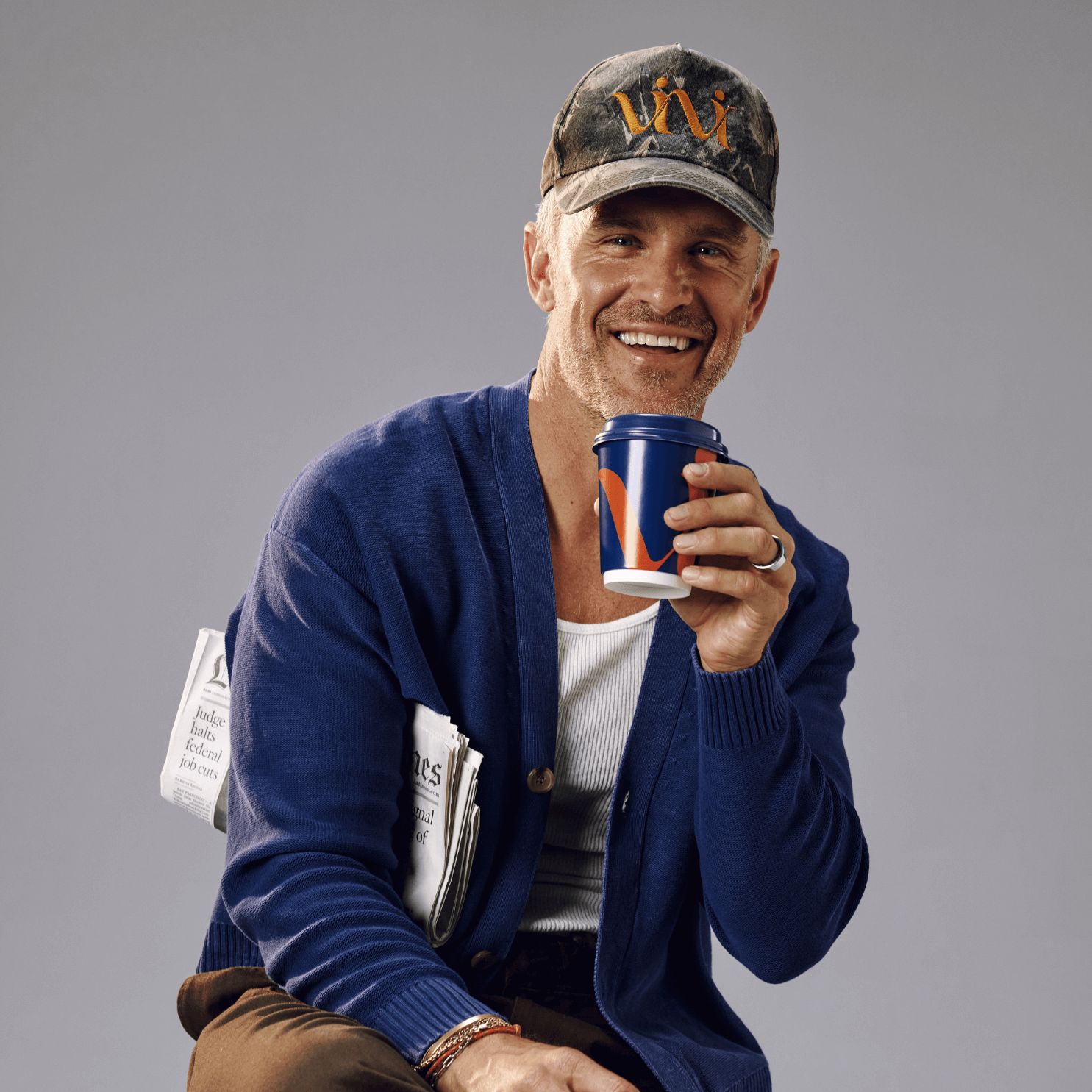 A smiling man in a cap and cardigan holds a coffee cup and a newspaper.