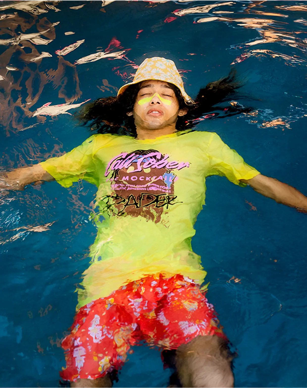 Person in colorful clothing floating in a pool wearing a yellow floppy hat.