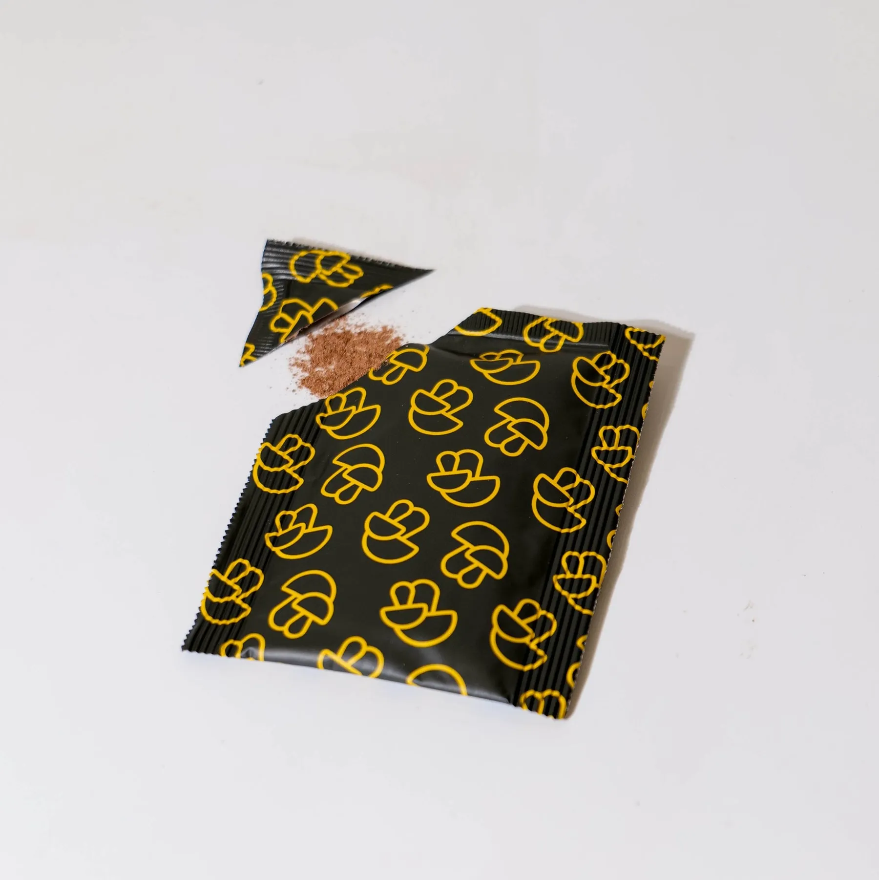 A black packet with a yellow mushroom pattern, torn open with brown powder spilling out.