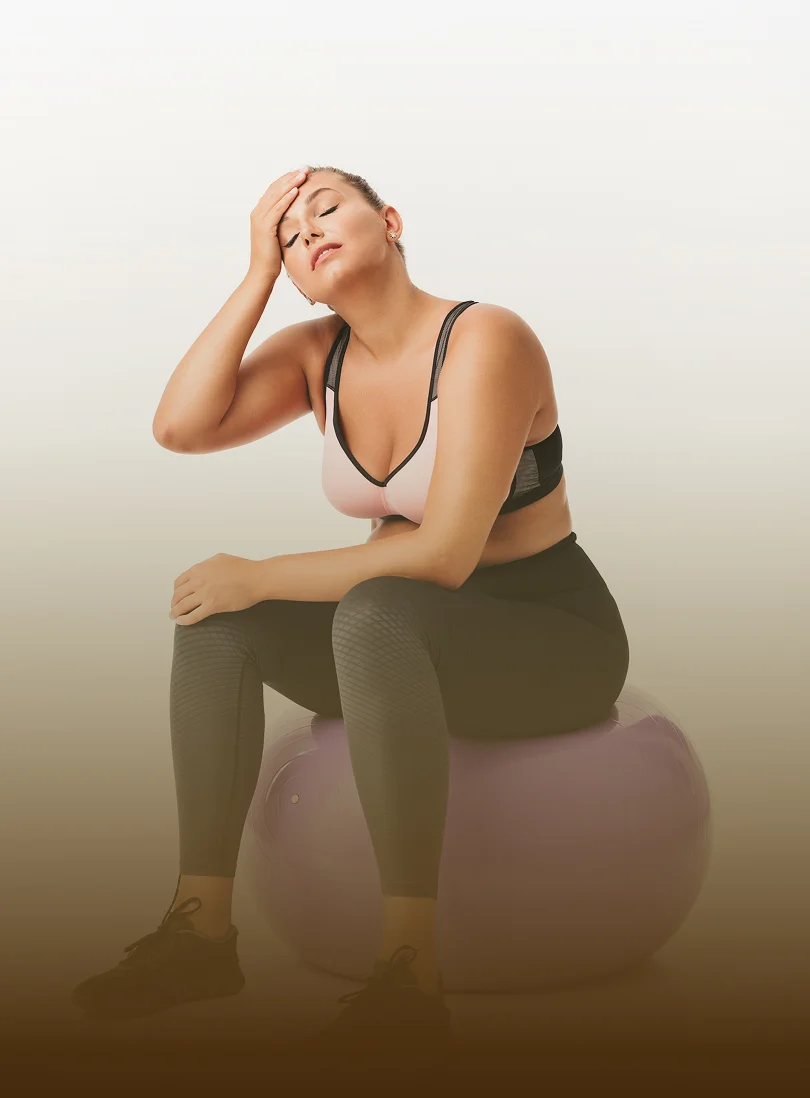 A woman in workout clothes sits on an exercise ball, resting with her hand on her forehead.