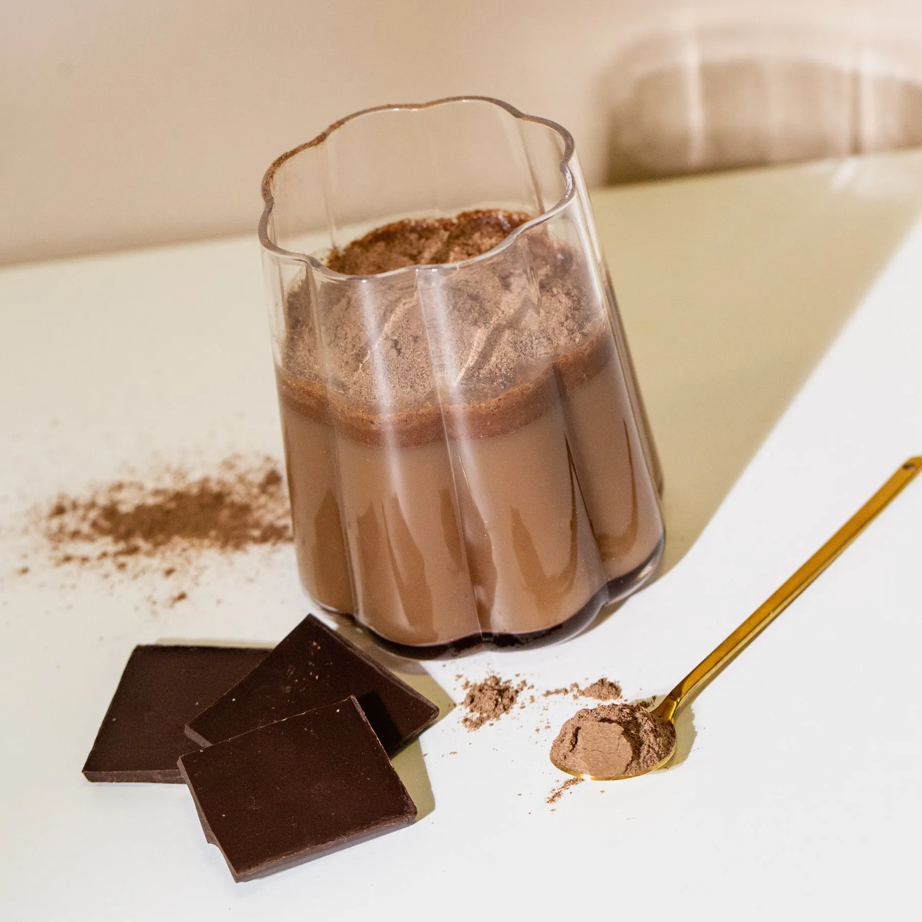 A glass of chocolate milk with cocoa powder, chocolate pieces, and a golden spoon on a white surface.