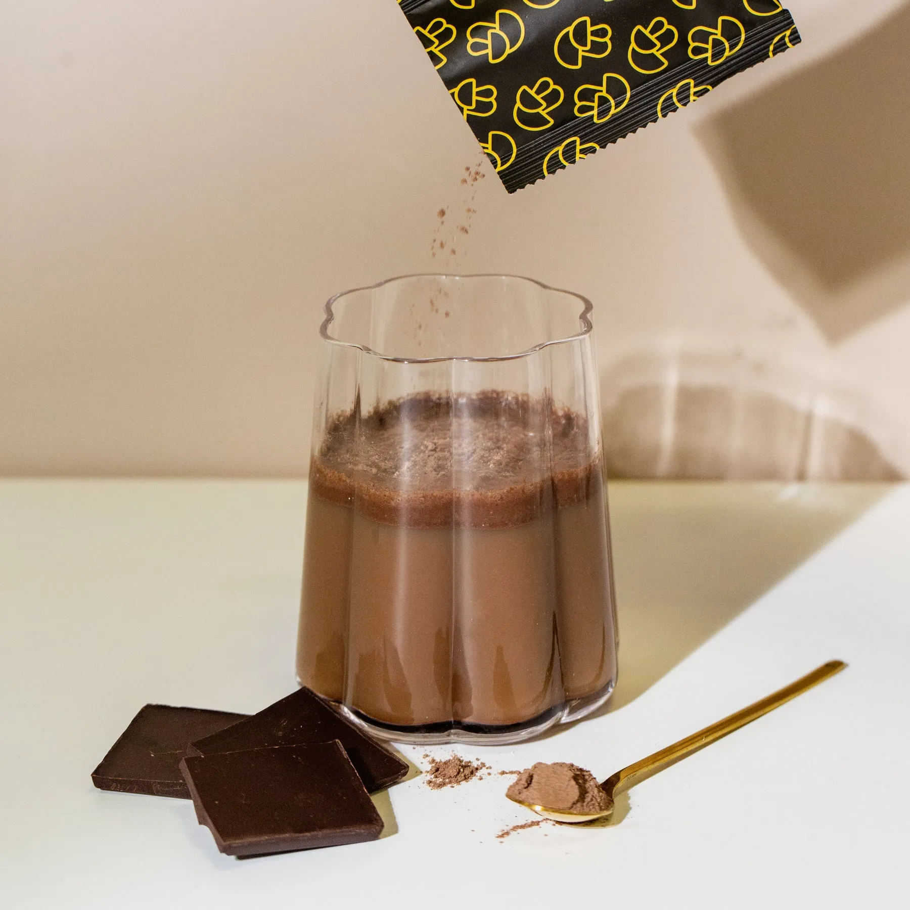 Powder from a packet is poured into a glass of chocolate drink, next to chocolate squares and a spoon.