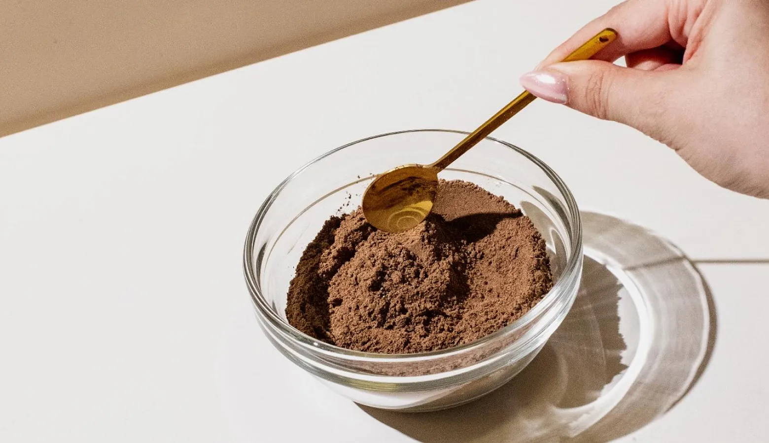 A hand dips a small golden spoon into a glass bowl of brown powder on a white surface.