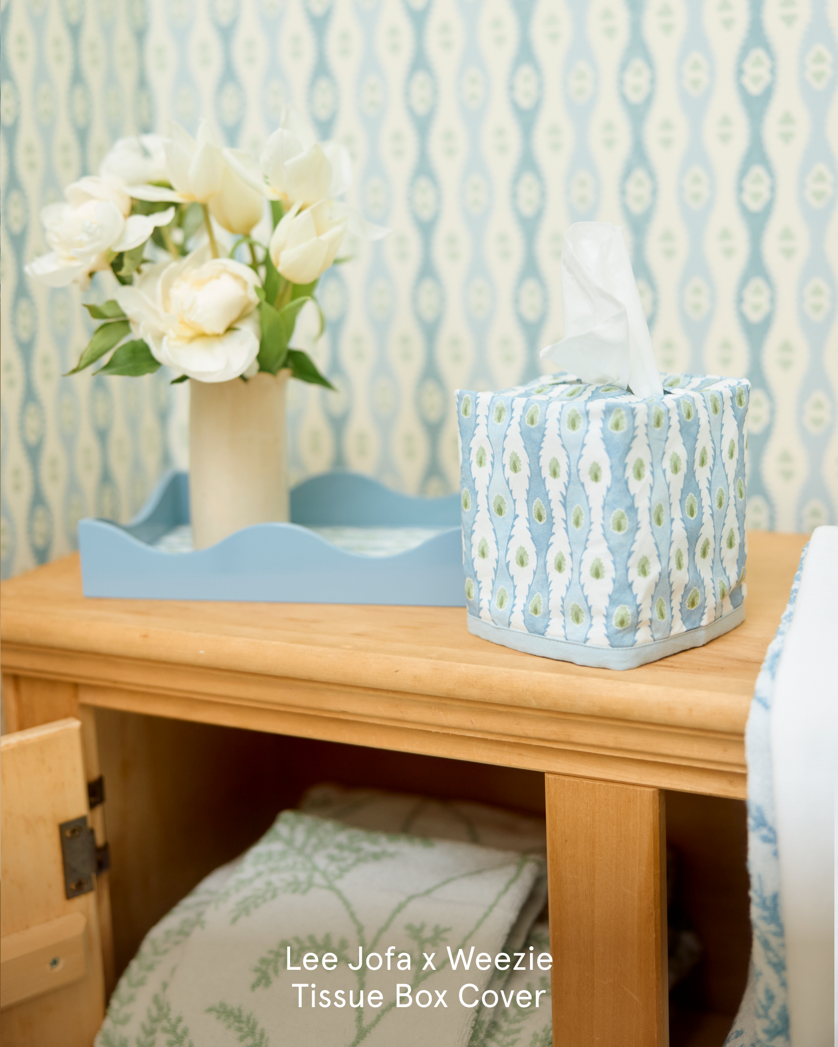 A patterned tissue box cover on a wooden nightstand next to a vase of white flowers.