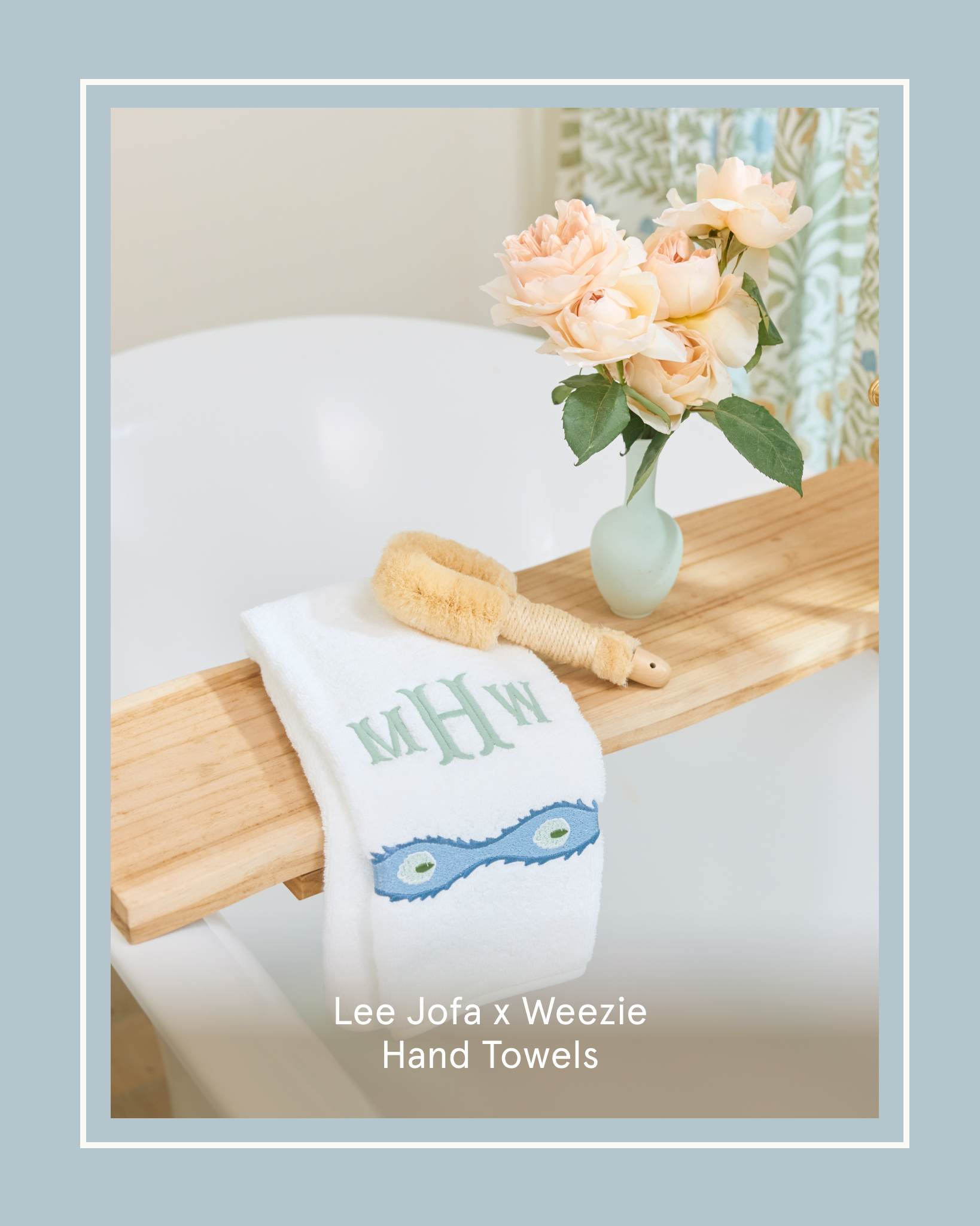A monogrammed hand towel, dry brush, and vase of roses on a wooden bath tray over a bathtub.