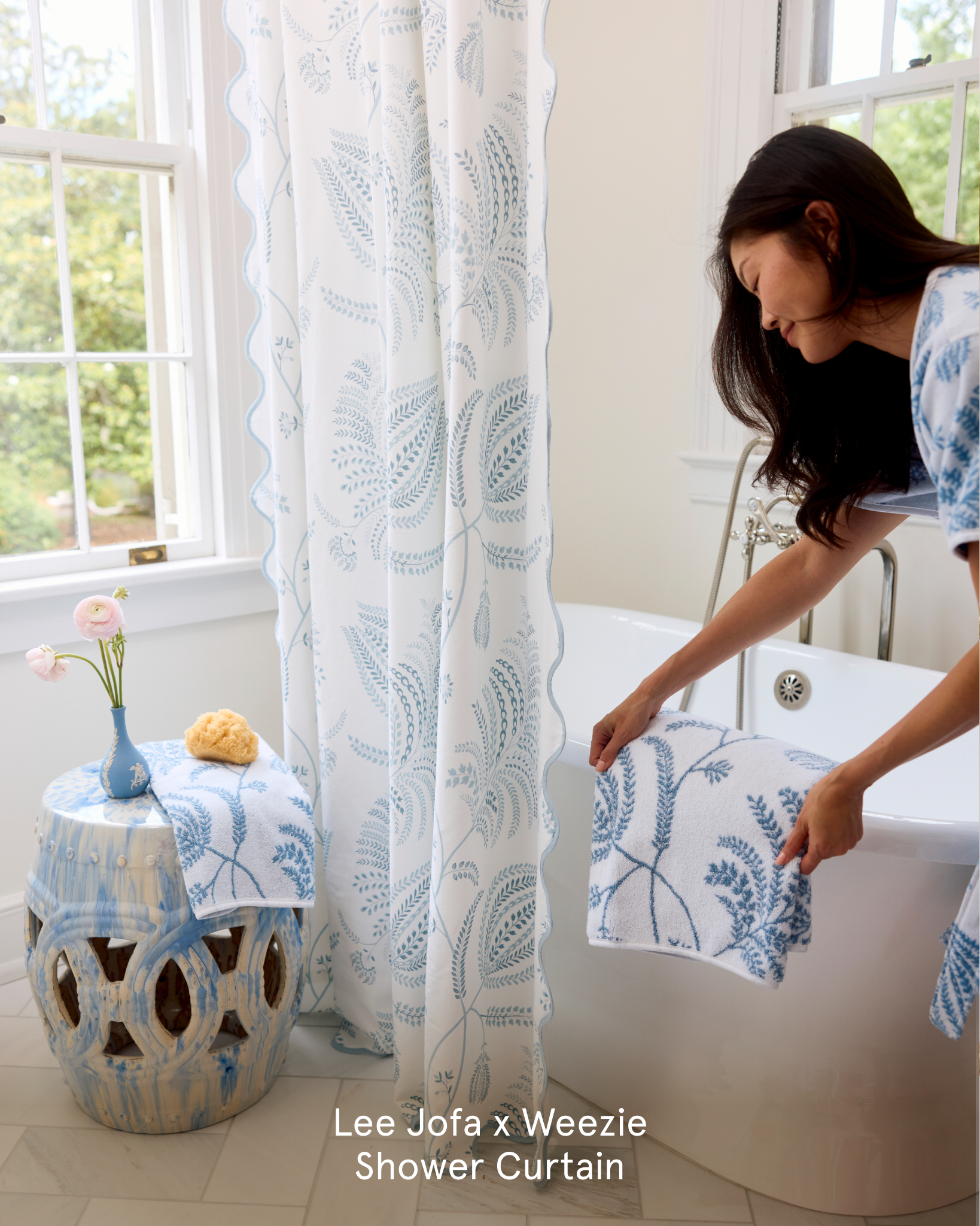 A woman places a towel with a blue botanical print on a bathtub beside a matching shower curtain.