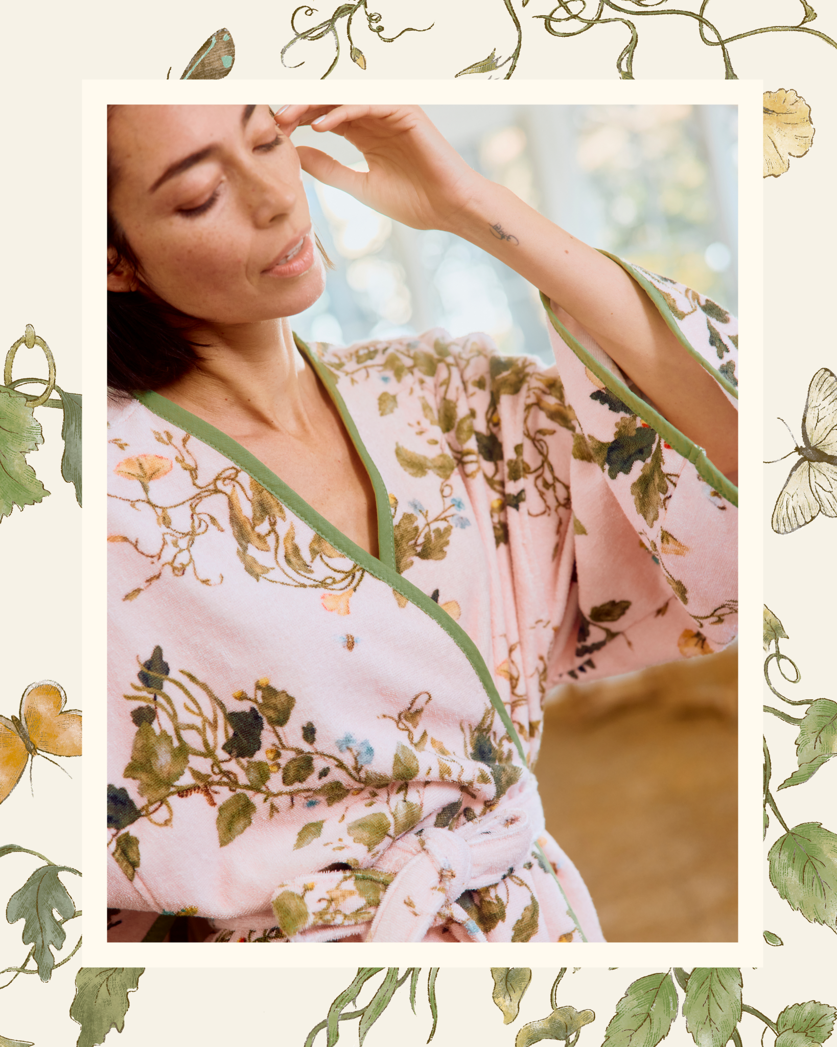 A woman with her eyes closed wears a pink floral robe with green trim, touching her temple.