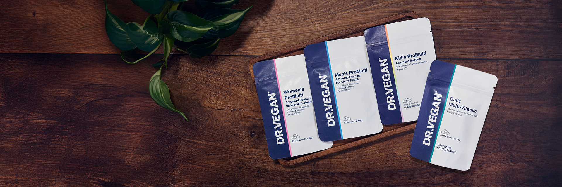 Four DR.VEGAN supplement packets on a wooden surface with a plant.