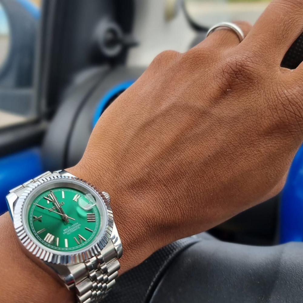 A hand on a steering wheel wearing a green-faced luxury watch.