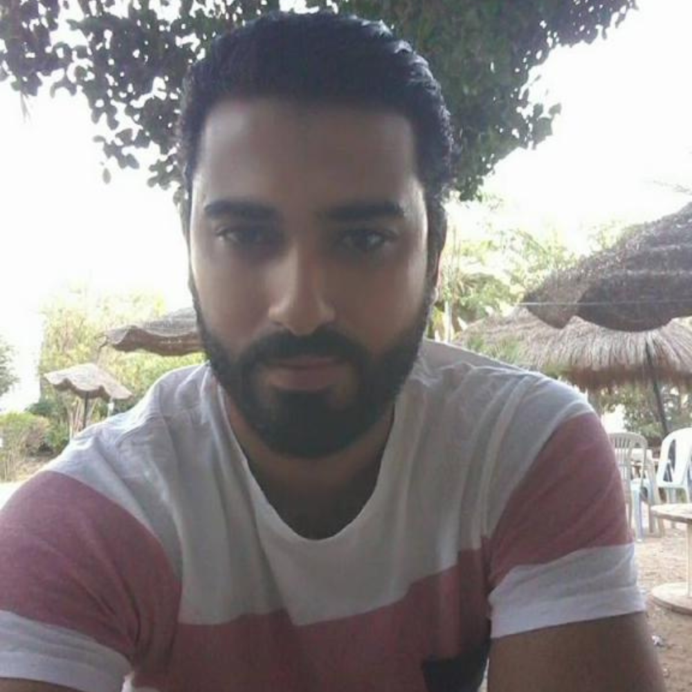 Man sitting outdoors with trees and straw umbrellas in the background.