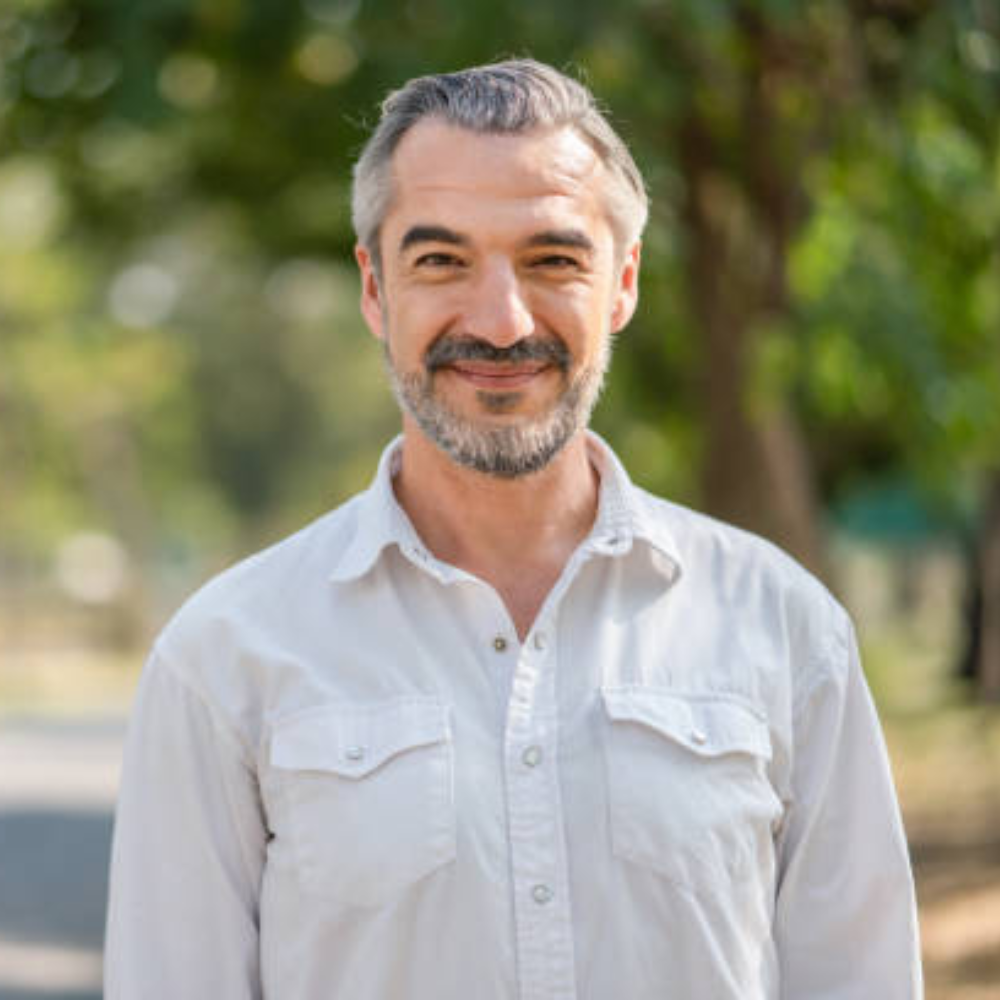 A man smiling outdoors in a white shirt with a blurred green background.