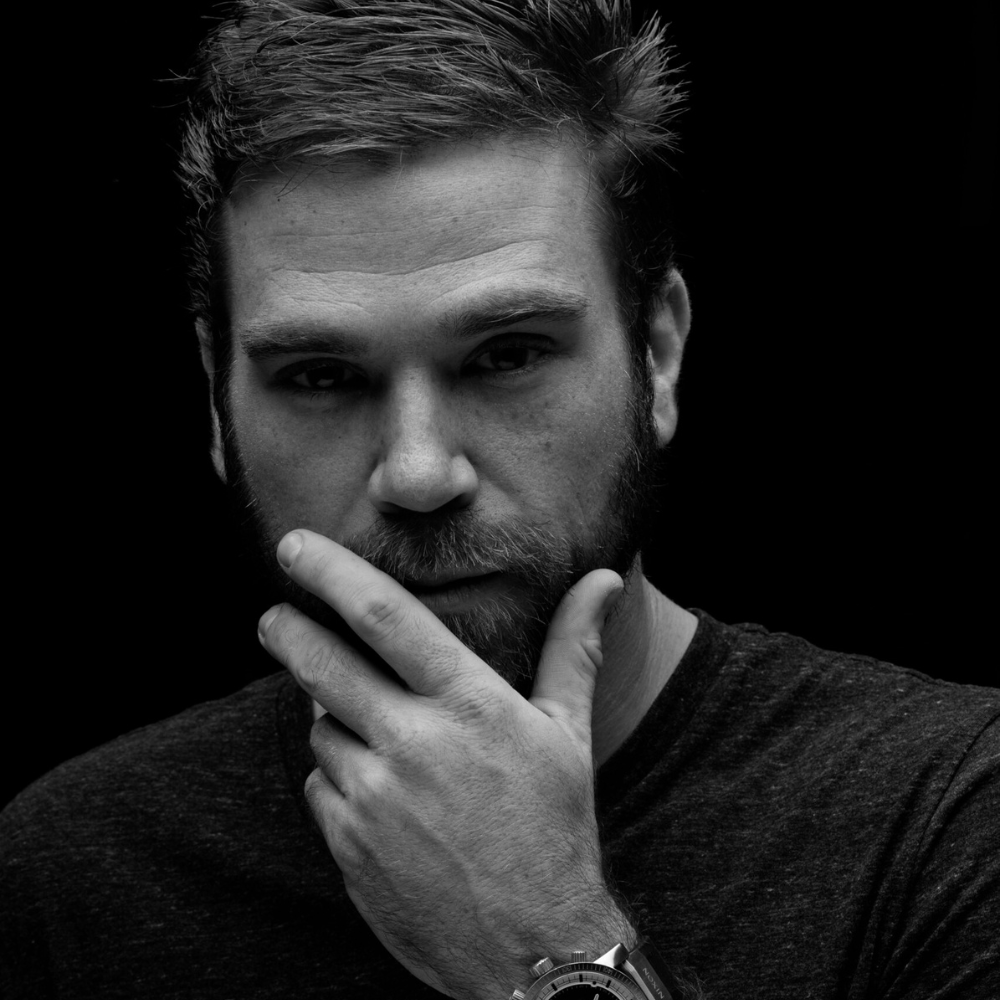A man with a beard thoughtfully touching his face, wearing a wristwatch, against a dark background.