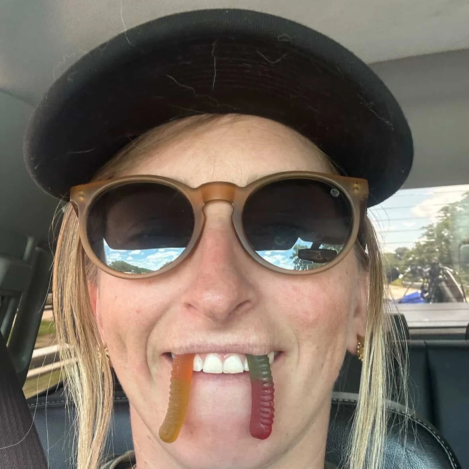 A woman wearing sunglasses and a hat smiles with two gummy worms hanging from her mouth.