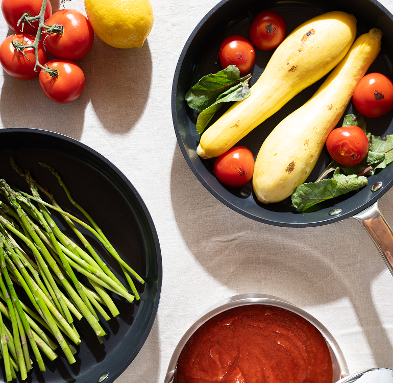 Fresh vegetables and tomato sauce arranged artfully on a table with two pans.