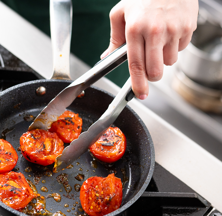 Hand using tongs to cook halved tomatoes in a frying pan.