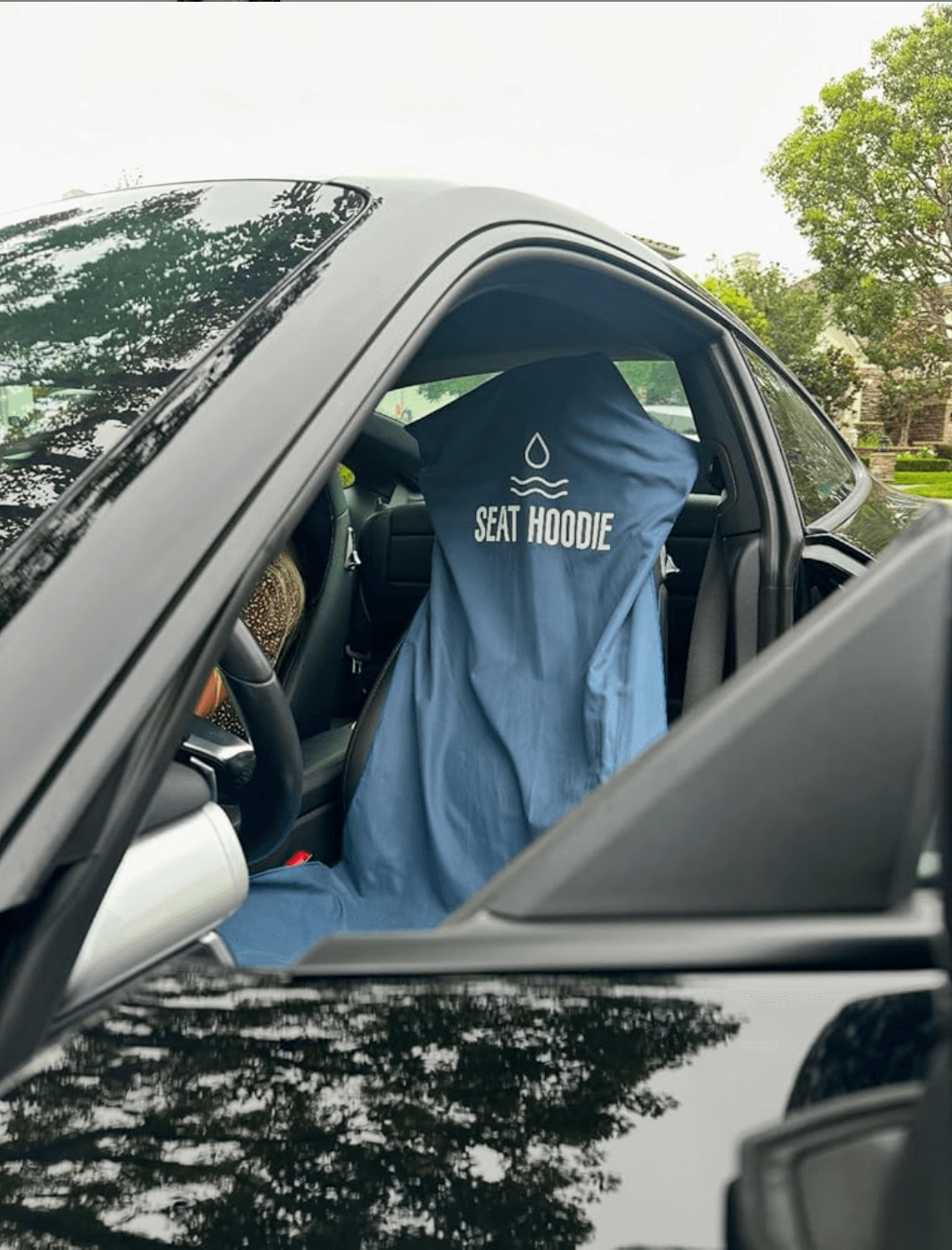 A blue 'Seat Hoodie' car seat cover is draped over the driver's seat of a black car.