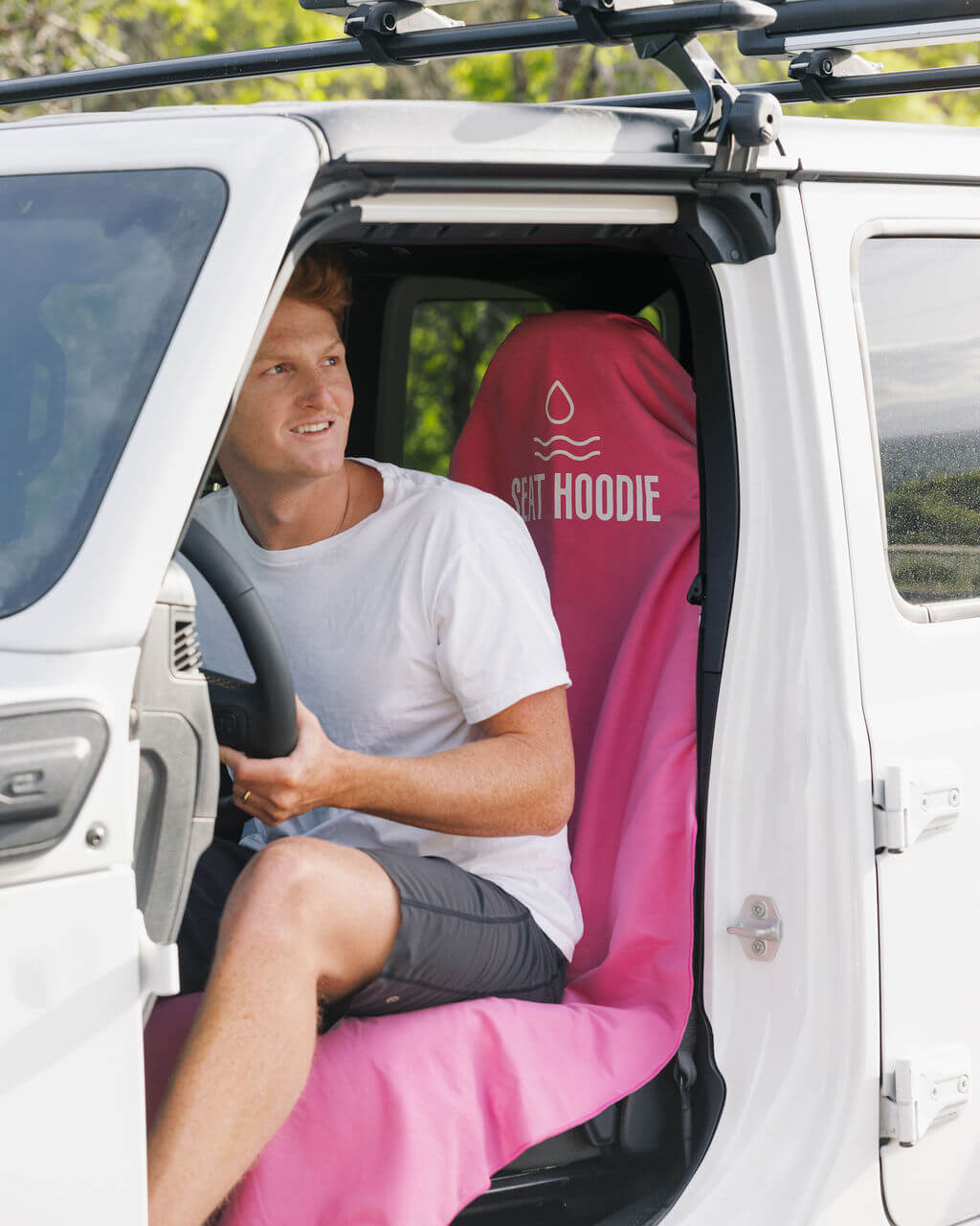 A man sits in the driver's seat of a white vehicle on a pink 'Seat Hoodie' car seat cover.