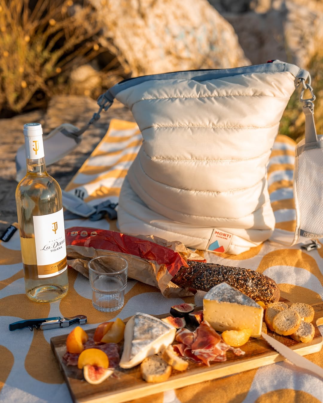 Picnic setup with wine, cheese, bread, and a bag on a patterned blanket.